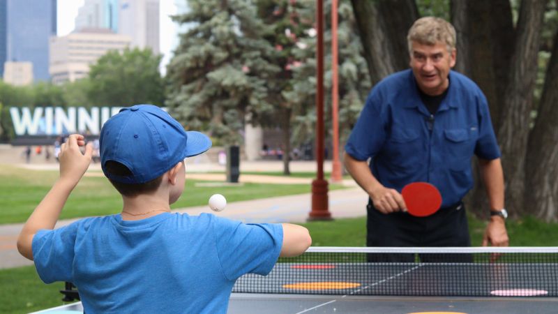 An older man and child playing ping pong outside at The Forks. 